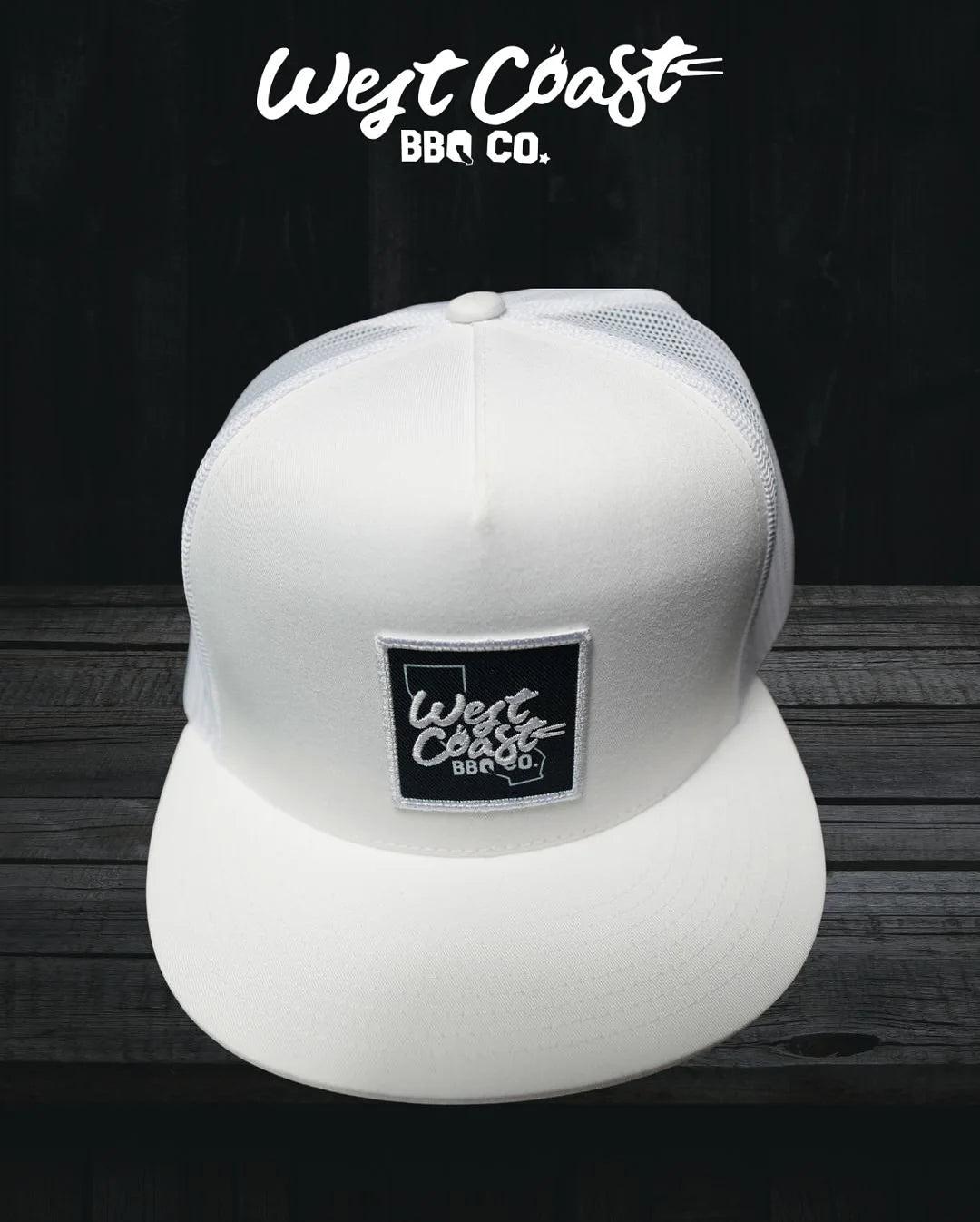 A white trucker hat with a black square patch featuring the "West Coast BBQ Co." logo, shown sitting on a dark wooden surface.