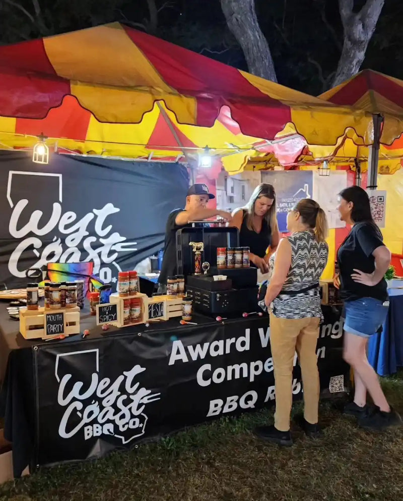 West Coast BBQ Co. booth at an outdoor event with people interacting.