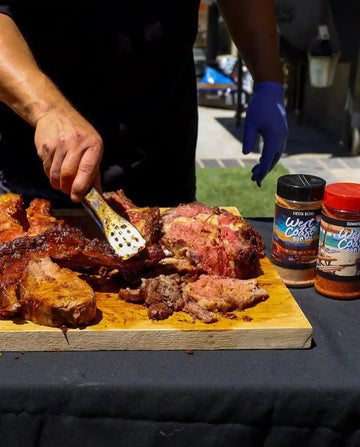 Person cutting into a large piece of meat on a wooden cutting board with two jars of seasoning in the background.